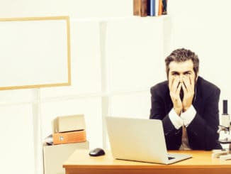 Tired young bearded man sitting at desk in front of computer with his chin resting on his hands at workplace