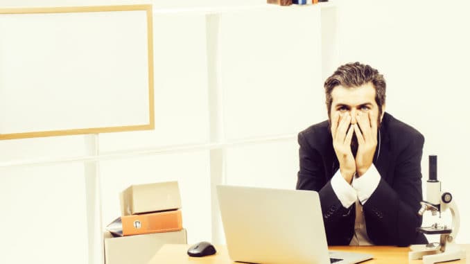 Tired young bearded man sitting at desk in front of computer with his chin resting on his hands at workplace