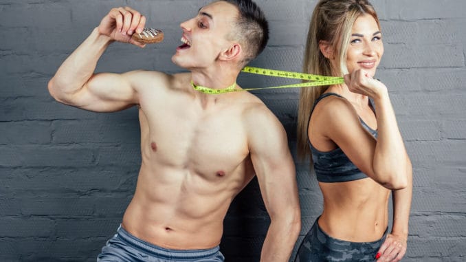 Beautiful blonde women in camouflage sports suit inflated and beautiful men with a measuring tape in the gym eating an orange and biscuits.