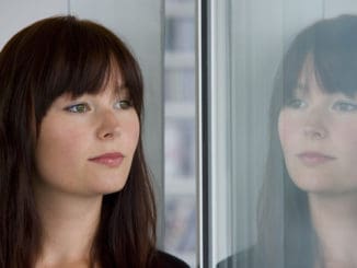 A beautiful young woman stares wistfully into a window where her reflection looks back at her.