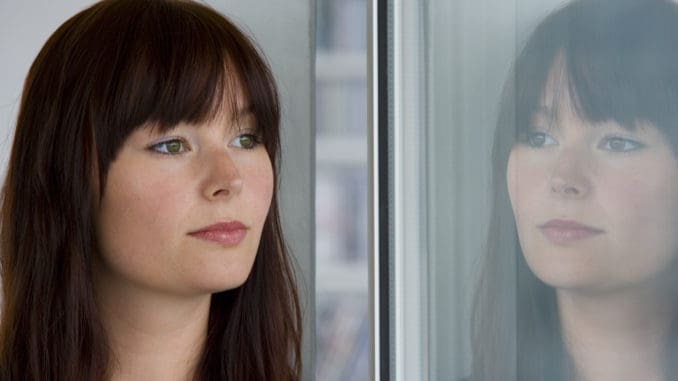 A beautiful young woman stares wistfully into a window where her reflection looks back at her.