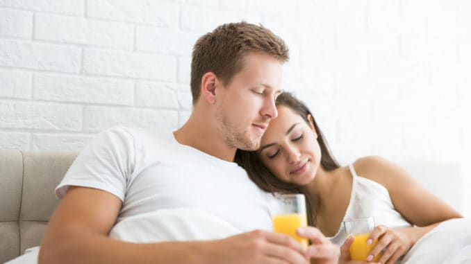 Happy young couple handsome man and beautiful woman lying in bed in bright bedroom in the morning drinking orange juice