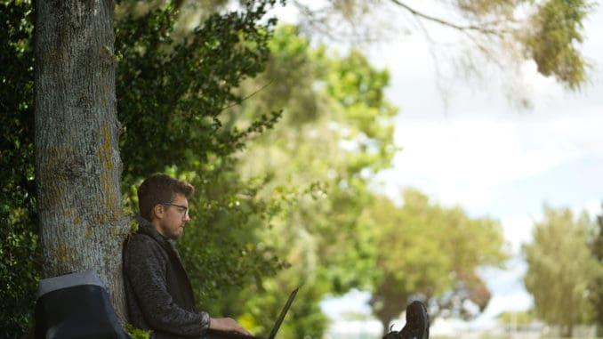 dreamstime_m_165410878 Man sitting under a tree, reposing and working on his computer.