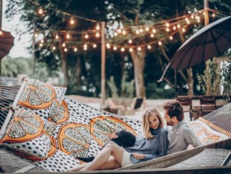 Stylish hipster couple cuddling and relaxing in hammock under retro lights in evening summer park.