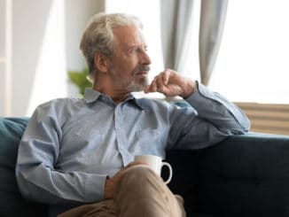 Pensive elderly 60s man sit relax on couch in living room drinking tea look in window distance thinking, thoughtful mature 50s husband rest on sofa at home feel sad pondering considering future