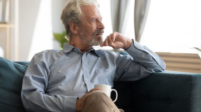 Pensive elderly 60s man sit relax on couch in living room drinking tea look in window distance thinking, thoughtful mature 50s husband rest on sofa at home feel sad pondering considering future