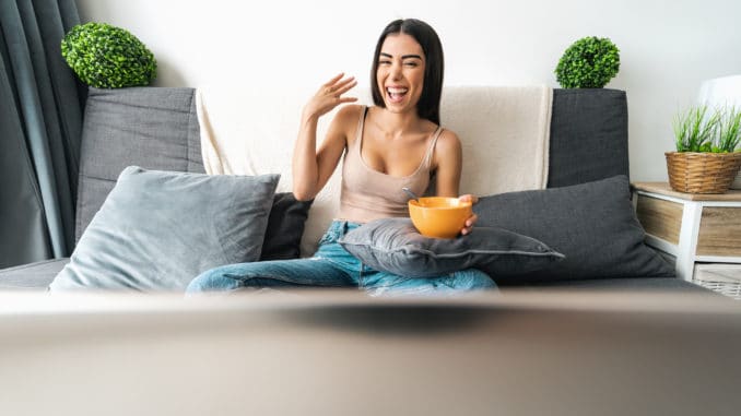 Happy woman eating cereal cup with milk while watching on computer laptop sitting on sofa - Health meal and living room entertainment concept