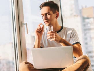 Portrait of young ill man taking pill and drink water while sitting with laptop on laps near window.