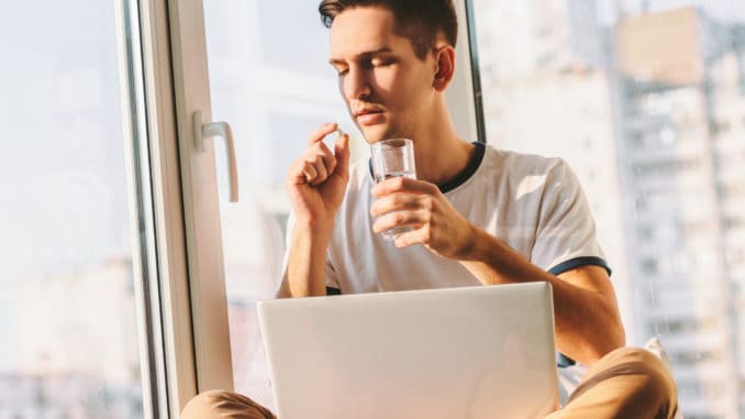 Portrait of young ill man taking pill and drink water while sitting with laptop on laps near window.