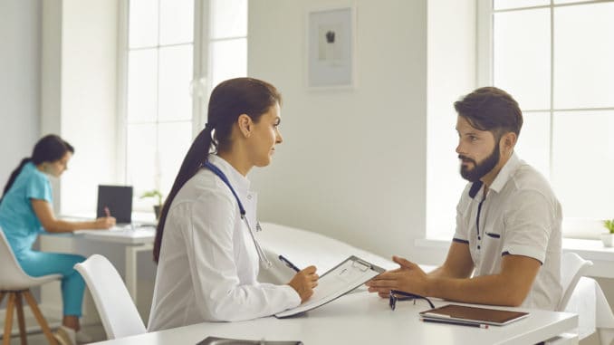 Medical clinic offfice. Young smiling woman doctor in medical uniform sitting and listening to talking man patient during consultation. Visiting doctor in hospital concept