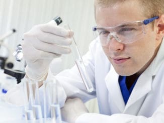 A male medical or scientific researcher or doctor looking at a test tube of clear liquid in a laboratory with microscopes.