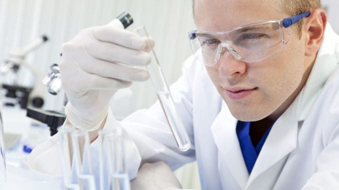 A male medical or scientific researcher or doctor looking at a test tube of clear liquid in a laboratory with microscopes.