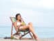 A young and attractive brunette Caucasian woman in a white swimsuit eating fruits and relaxing. The image is taken on a beach background.