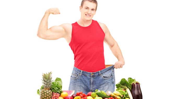 Weight loss man showing his muscles and standing behind a pile o Weight loss man showing his muscles and standing behind a pile of fruits and vegetables, isolated on white background