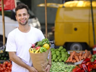 Man carrying a shopping paper bag full of fruits and vegetables at an open street market.