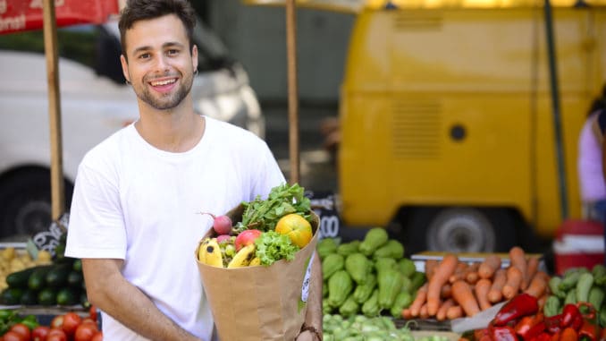 Man carrying a shopping paper bag full of fruits and vegetables at an open street market.