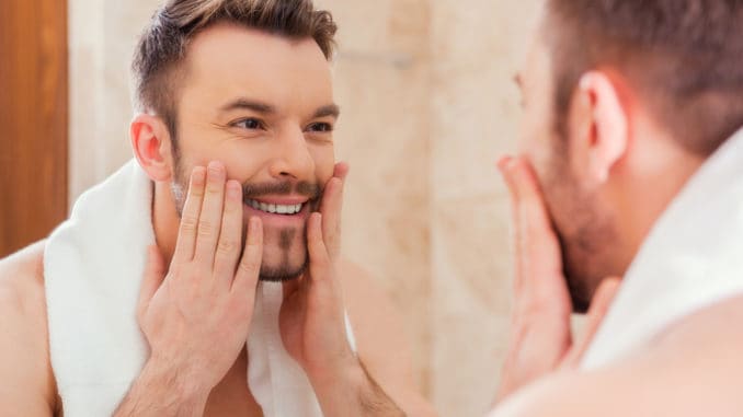 dreamstime_m_49634205 Handsome young man touching his face and smiling while standing in front of the mirror