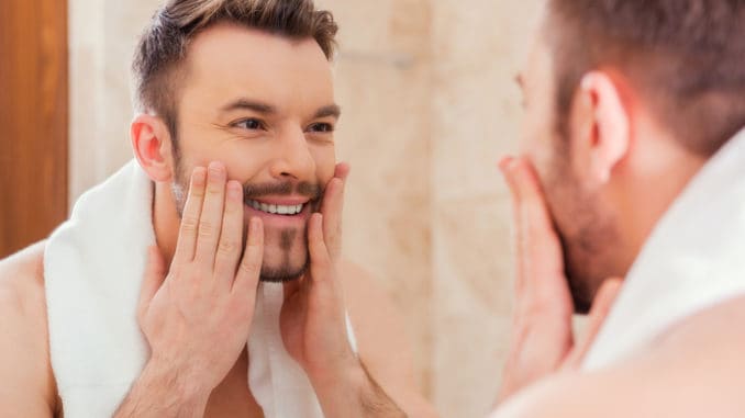 Handsome young man touching his face and smiling while standing in front of the mirror