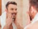 Handsome young man touching his face and smiling while standing in front of the mirror