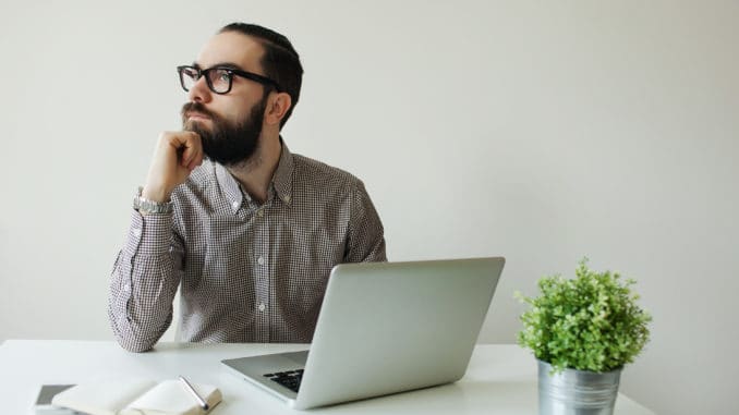 Busy man with beard in glasses thinking over laptop with smartphone on the table