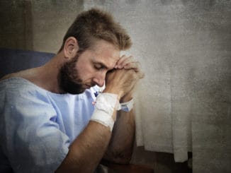 Young injured man in hospital room sitting alone in pain looking negative and worried for his bad health condition sitting on chair suffering depression on a sad lonely background