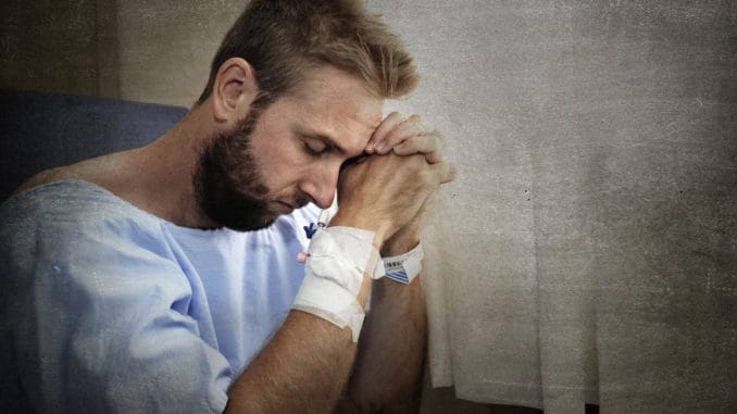 Young injured man in hospital room sitting alone in pain looking negative and worried for his bad health condition sitting on chair suffering depression on a sad lonely background