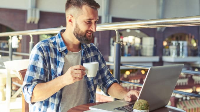 Young adult man drinking coffee and using laptop in cafe