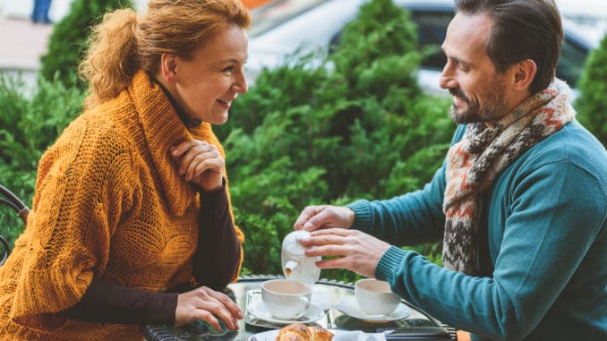 Happy married couple is having breakfast together. They are sitting at table on street and smiling. Man is pouring tea into cup