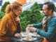 Happy married couple is having breakfast together. They are sitting at table on street and smiling. Man is pouring tea into cup