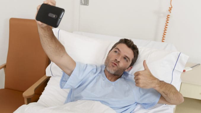 Young attractive man lying on bed hospital clinic holding mobile phone taking self portrait selfie photo giving thumb up in optimist recovery concept and health care positive image