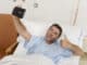 Young attractive man lying on bed hospital clinic holding mobile phone taking self portrait selfie photo giving thumb up in optimist recovery concept and health care positive image