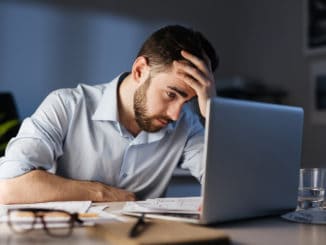 Portrait of tired bearded man working overtime alone in dark office late at night, using laptop and resting head on hand looking at screen