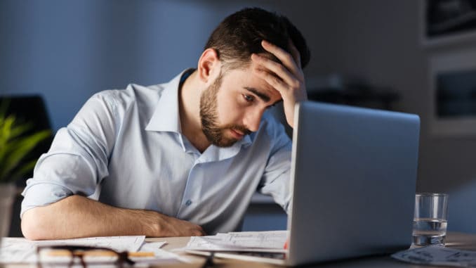 Portrait of tired bearded man working overtime alone in dark office late at night, using laptop and resting head on hand looking at screen