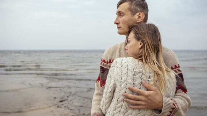 Happy thoughtful couple standing on a rock beach near sea hugging each other in cold foggy cloudy autumn weather. Copy space