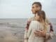 Happy thoughtful couple standing on a rock beach near sea hugging each other in cold foggy cloudy autumn weather. Copy space