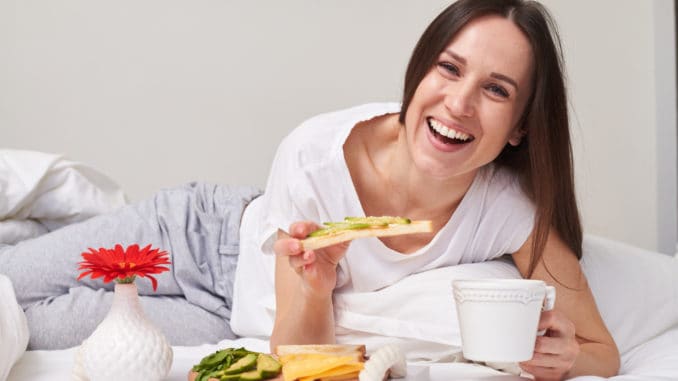 Mid shot of beautiful smiling girl eating sandwich with avocado and drinking aromatic tea