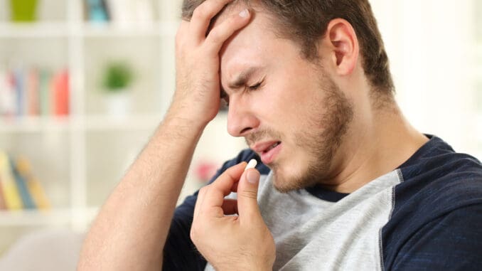 Man suffering headache taking a pill sitting on a sofa in the living room in a house interior