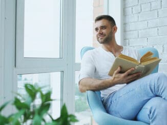 Young man reading book sitting on balcony in modern apartment