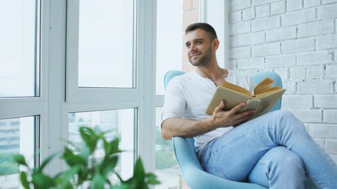 Young man reading book sitting on balcony in modern apartment