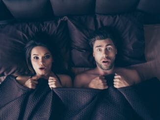 Funny lovers men and women lying in bed covered by sheets with wide opened eyes