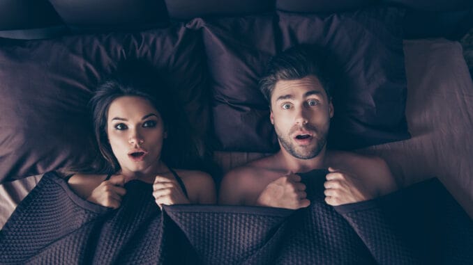 Funny lovers men and women lying in bed covered by sheets with wide opened eyes