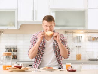 Young man eating tasty toasted bread at table in kitchen