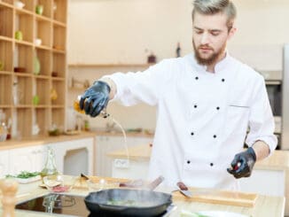 Portrait of handsome professional chef working in modern restaurant kitchen standing at wooden table and adding oils to delicious dishes