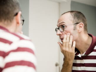 Reflective image of a man looking into a mirror as he touches his cheek and looks to the side