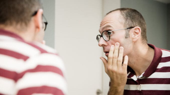 Reflective image of a man looking into a mirror as he touches his cheek and looks to the side