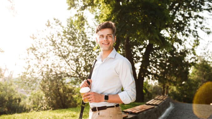 Happy young man in shirt walking at the city park with cup of coffee