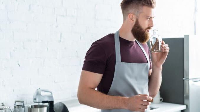 dreamstime_s_128980246 handsome bearded young man in apron holding container with cinnamon sticks in kitchen