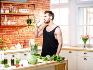 Handsome sports man in black t-shirt drinking milk shake standing on the kitchen full of healthy food at home