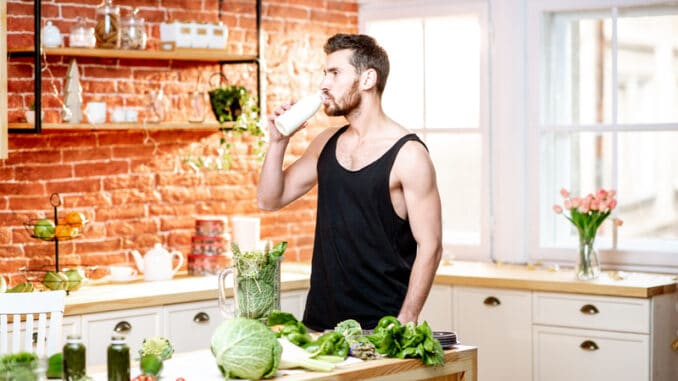 Handsome sports man in black t-shirt drinking milk shake standing on the kitchen full of healthy food at home