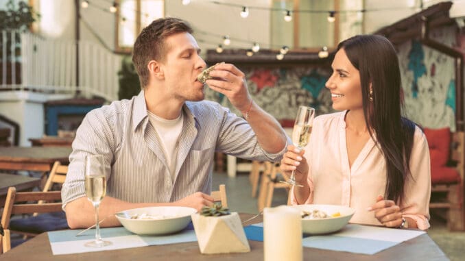 Drinking sparkling wine. Light-haired boyfriend with stubble eating oyster directly from hard shell during lunch with his girl